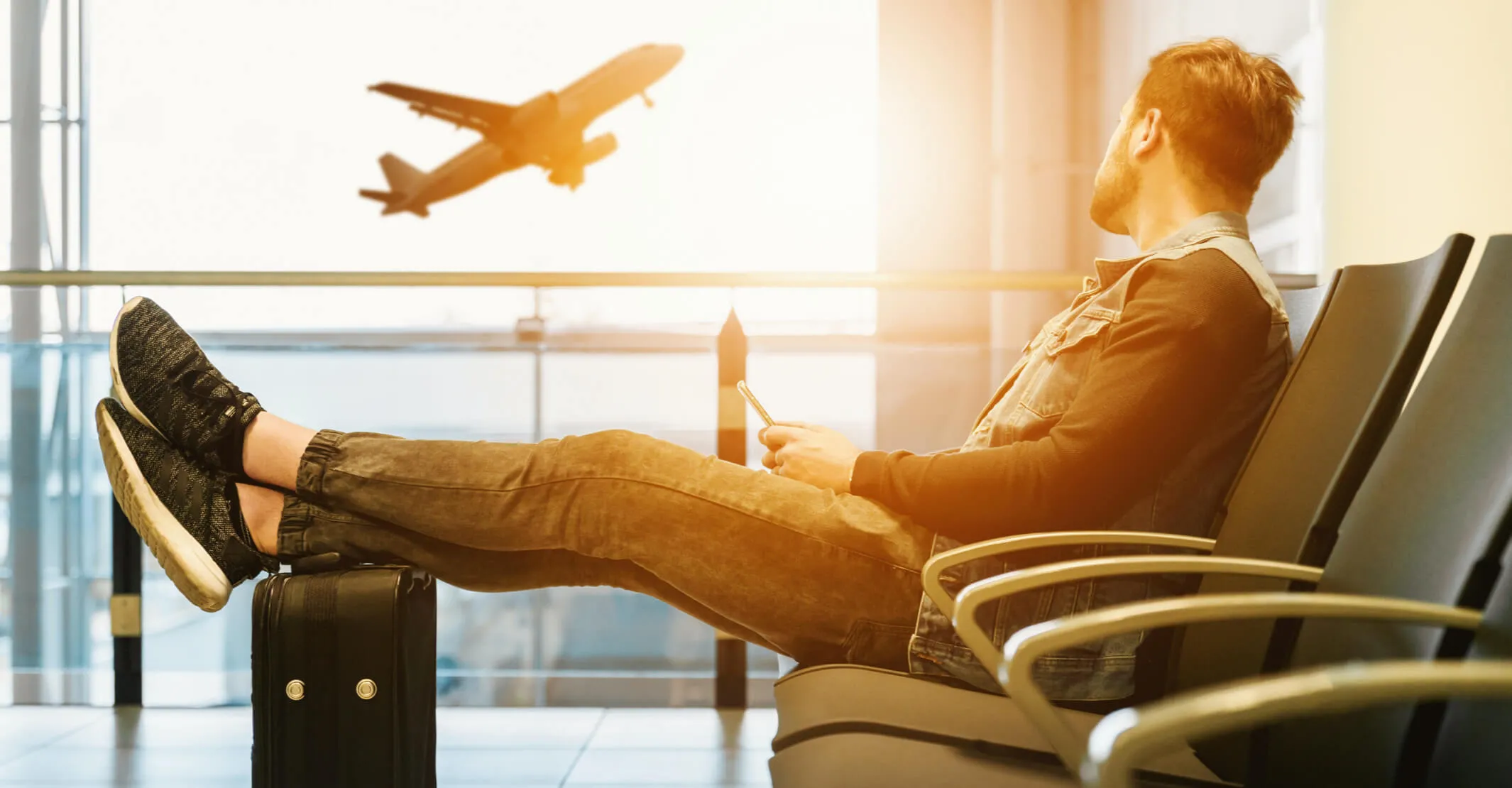 man sitting in airport with his feet up on his luggage watching plane take off